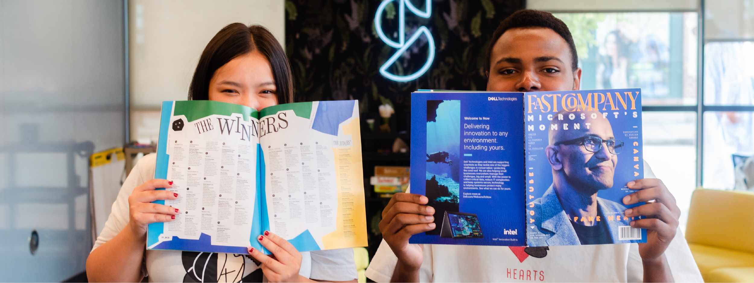 Two students in Shift Retail Lab at VCU, holding up the Fast Company 2024 Award Winners Magazine, in which Shift Retail Lab is listed. One students holds the magazine open to the page of winners while the other reads the magazine.